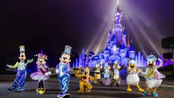 Mickey Mouse et ses amis vêtus de costumes colorés devant le château de Cendrillon à Disneyland Paris, éclairé par des projecteurs sur fond de brume lors de leur célébration d'anniversaire.