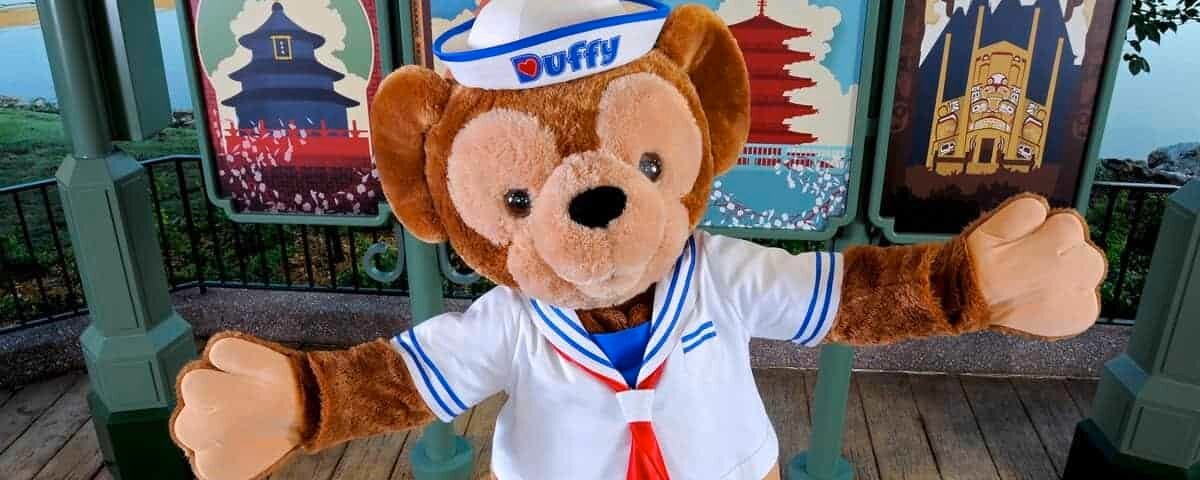 Un ours Duffy portant une tenue de marin avec un chapeau et une écharpe rouge se tient devant une exposition de panneaux d'affiches colorés sur le thème de pays, notamment la Chine, le Mexique, le Japon et le Canada.