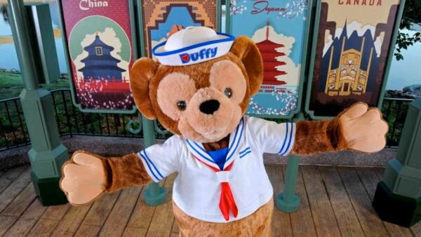 Un ours Duffy portant une tenue de marin avec un chapeau et une écharpe rouge se tient devant une exposition de panneaux d'affiches colorés sur le thème de pays, notamment la Chine, le Mexique, le Japon et le Canada.