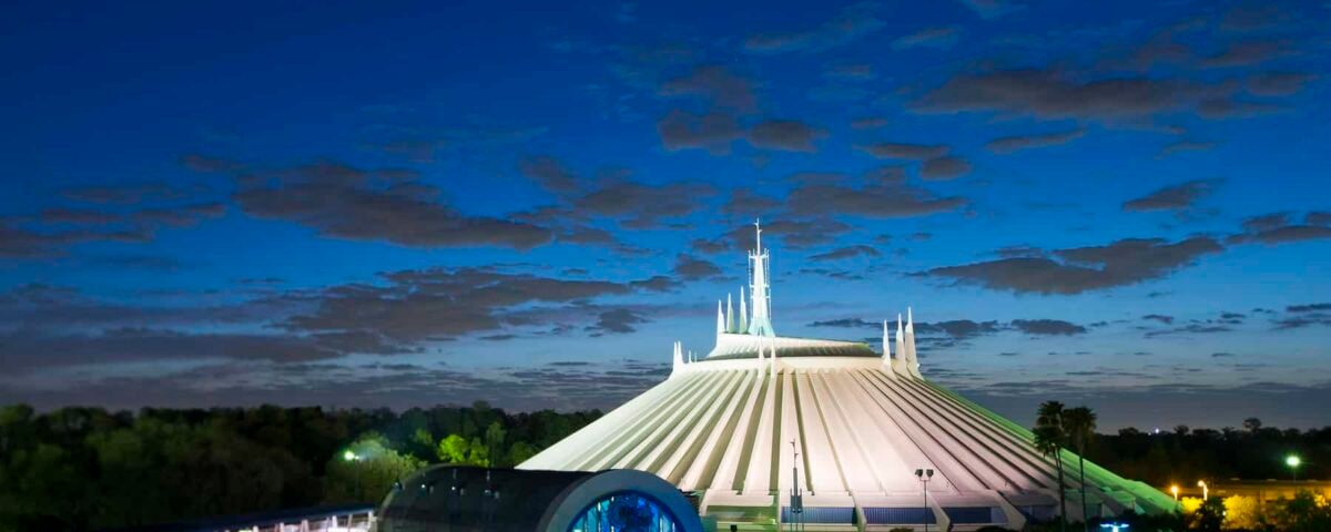 Vue nocturne de Space Mountain avec un toit blanc en forme de tente et de multiples flèches, illuminé par un ciel crépusculaire, entouré d'une verdure luxuriante et d'un sentier éclairé.