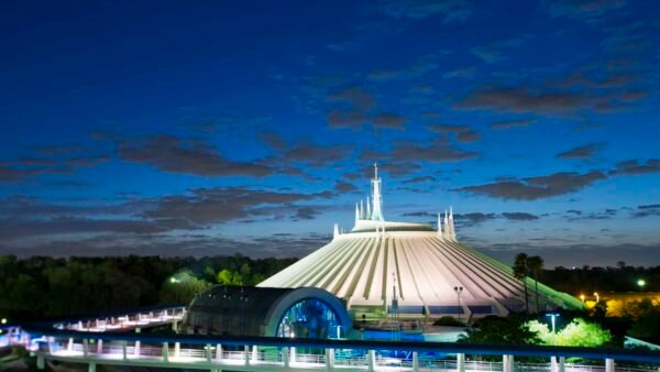 Vue nocturne de Space Mountain avec un toit blanc en forme de tente et de multiples flèches, illuminé par un ciel crépusculaire, entouré d'une verdure luxuriante et d'un sentier éclairé.