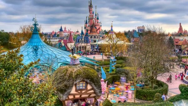 Une scène vibrante de Disneyland Paris, avec des bâtiments colorés, des foules animées à Fantasyland et l'emblématique Château de la Belle au Bois Dormant en arrière-plan sous un ciel nuageux.