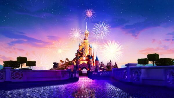 Des feux d'artifice illuminent le ciel au-dessus de Disneyland Paris au crépuscule, avec un reflet scintillant dans l'eau des douves environnantes.