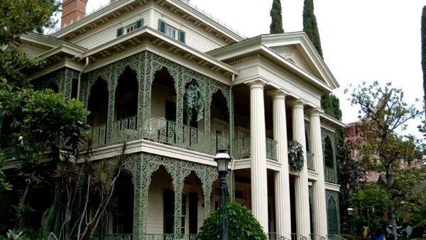 Un grand manoir orné avec une conception architecturale classique, connu localement sous le nom de The Haunted Mansion, avec de hauts piliers, des balustrades de balcon en fer complexes et entouré d'une verdure luxuriante.