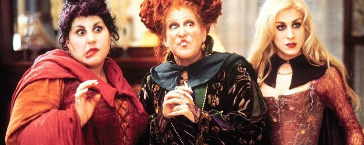 Trois personnages sorciers du film "Hocus Pocus" se tenant ensemble dans une pièce, l'air surpris. Les sorcières portent des costumes élaborés et colorés avec des coiffures uniques.