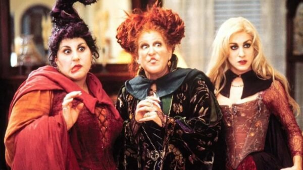 Trois personnages sorciers du film "Hocus Pocus" se tenant ensemble dans une pièce, l'air surpris. Les sorcières portent des costumes élaborés et colorés avec des coiffures uniques.