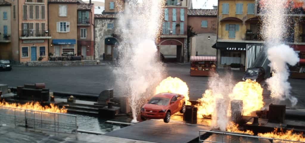 Une voiture rouge effectue une cascade entourée de feu et d'eau qui éclate dans un décor extérieur qui ressemble à une place de village européenne pittoresque, avec les lumières du spectacle Motors Action.