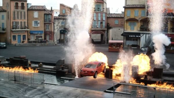Une voiture rouge effectue une cascade entourée de feu et d'eau qui éclate dans un décor extérieur qui ressemble à une place de village européenne pittoresque, avec les lumières du spectacle Motors Action.