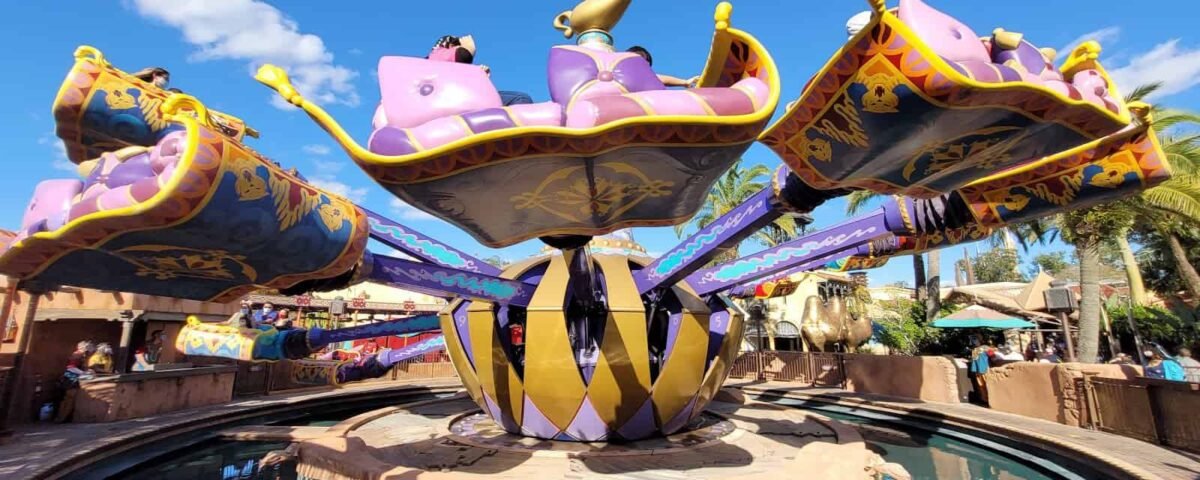 Un manège coloré et fantaisiste ressemblant aux tapis magiques d'Aladdin avec des accents dorés et violets, entouré d'un étang d'eau réfléchissant sous un ciel bleu vif avec peu de nuages.