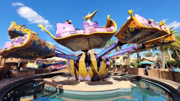 Un manège coloré et fantaisiste ressemblant aux tapis magiques d'Aladdin avec des accents dorés et violets, entouré d'un étang d'eau réfléchissant sous un ciel bleu vif avec peu de nuages.