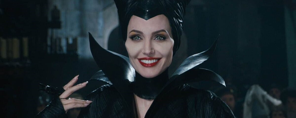 Une femme incarnant un personnage maléfique avec des cornes sombres et stylisées et un costume noir sourit énigmatiquement dans un décor atmosphérique faiblement éclairé.