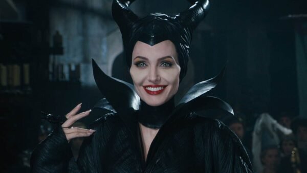 Une femme incarnant un personnage maléfique avec des cornes sombres et stylisées et un costume noir sourit énigmatiquement dans un décor atmosphérique faiblement éclairé.