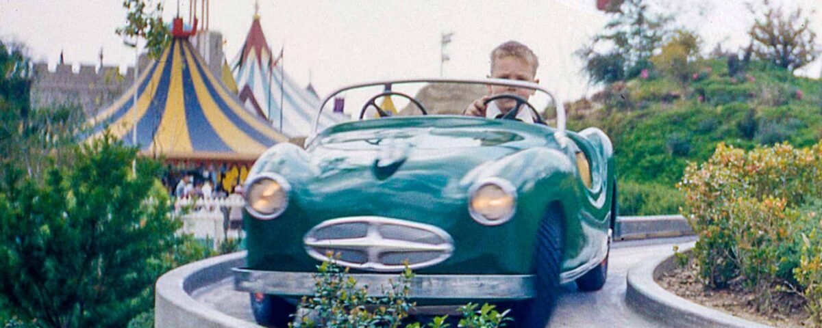 Un jeune garçon conduit une voiture verte de style vintage sur une piste guidée dans un parc d'attractions, désigné comme Midget Autopia, avec une atmosphère de carnaval colorée en arrière-plan.