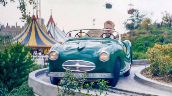 Un jeune garçon conduit une voiture verte de style vintage sur une piste guidée dans un parc d'attractions, désigné comme Midget Autopia, avec une atmosphère de carnaval colorée en arrière-plan.