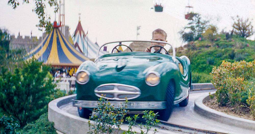 Un jeune garçon conduit une voiture verte de style vintage sur une piste guidée dans un parc d'attractions, désigné comme Midget Autopia, avec une atmosphère de carnaval colorée en arrière-plan.