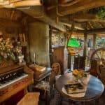 Chambre confortable et rustique avec un éclairage chaleureux, comprenant une table à manger pour deux, un piano vintage, des poutres en bois et une abondance de plantes vertes dans un style cabane familiale suisse