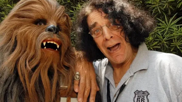 Un homme aux cheveux gris bouclés posant joyeusement à côté de Peter Mayhew dans un costume de Chewbacca, tous deux faisant des grimaces espiègles, sur fond de verdure.