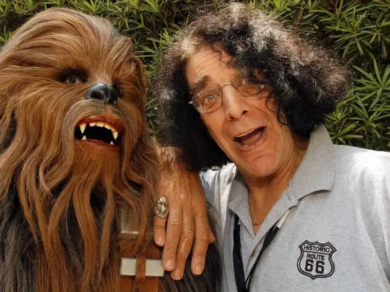 Un homme aux cheveux gris bouclés posant joyeusement à côté de Peter Mayhew dans un costume de Chewbacca, tous deux faisant des grimaces espiègles, sur fond de verdure.