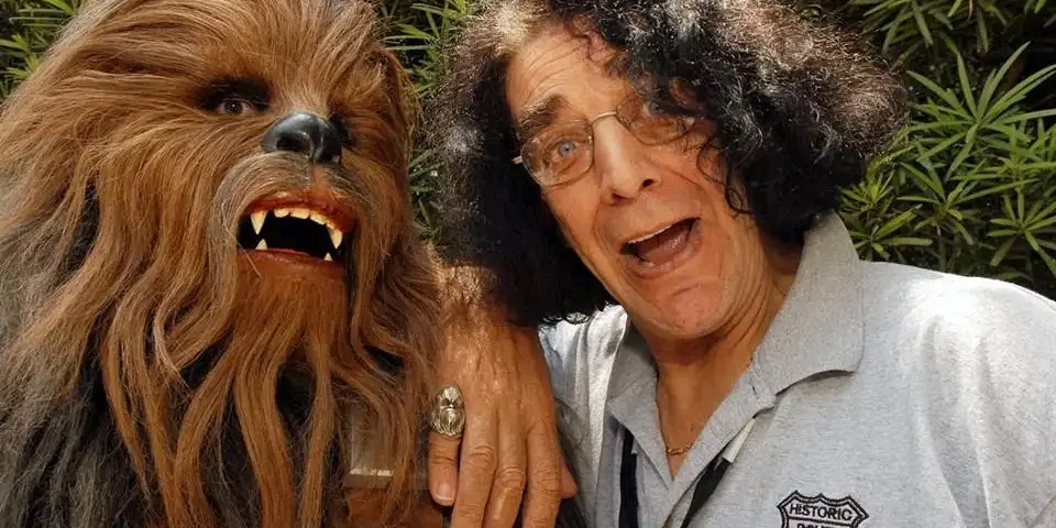 Un homme aux cheveux gris bouclés posant joyeusement à côté de Peter Mayhew dans un costume de Chewbacca, tous deux faisant des grimaces espiègles, sur fond de verdure.