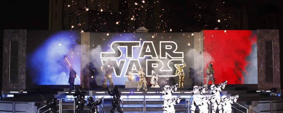 Un spectacle de festival vibrant mettant en vedette des personnages en costumes, notamment des stormtroopers et des pirates, sous une pancarte étincelante. Des fonds bleus et rouges complètent la scène.