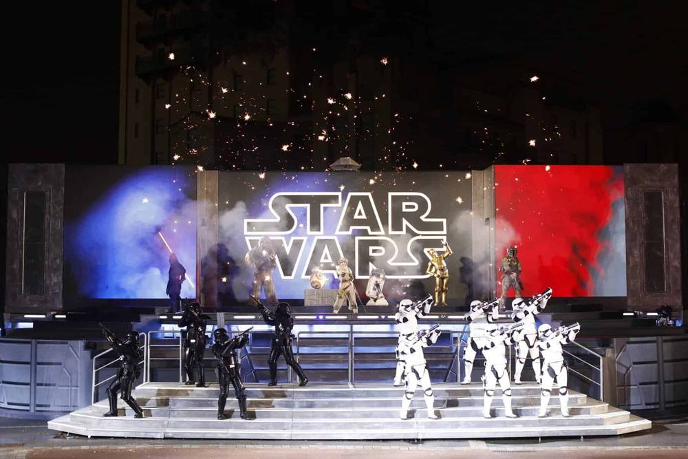 Un spectacle de festival vibrant mettant en vedette des personnages en costumes, notamment des stormtroopers et des pirates, sous une pancarte étincelante. Des fonds bleus et rouges complètent la scène.