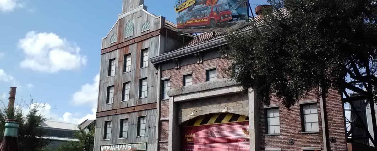 Une scène de rue dans un parc à thème dans la magie, avec des visiteurs marchant devant des bâtiments stylisés ressemblant à d'anciennes usines, ornés d'enseignes industrielles colorées, sous un ciel bleu clair.