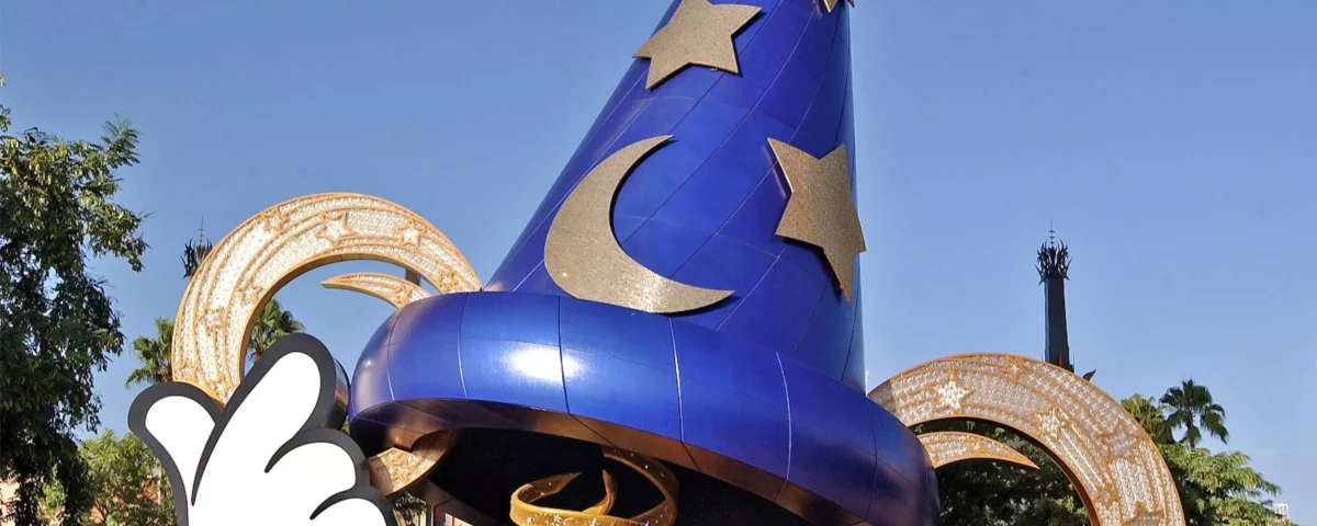 Un grand chapeau de sorcier bleu avec des motifs d’étoiles et de lune se dresse bien en évidence devant des bobines de film dorées, sous un ciel dégagé aux studios Disney MGM. Une main gantée de blanc, ressemblant à celle de Mickey Mouse