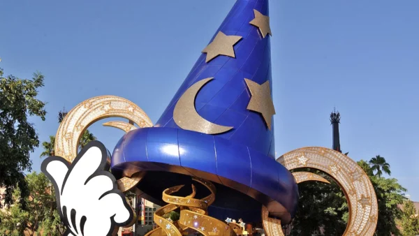 Un grand chapeau de sorcier bleu avec des motifs d’étoiles et de lune se dresse bien en évidence devant des bobines de film dorées, sous un ciel dégagé aux studios Disney MGM. Une main gantée de blanc, ressemblant à celle de Mickey Mouse