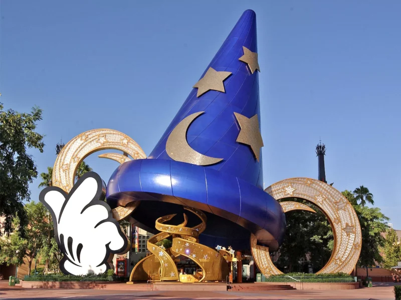 Un grand chapeau de sorcier bleu avec des motifs d’étoiles et de lune se dresse bien en évidence devant des bobines de film dorées, sous un ciel dégagé aux studios Disney MGM. Une main gantée de blanc, ressemblant à celle de Mickey Mouse