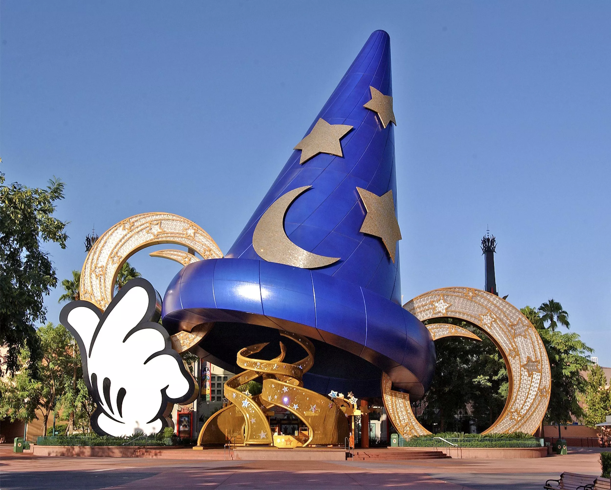 Un grand chapeau de sorcier bleu avec des motifs d’étoiles et de lune se dresse bien en évidence devant des bobines de film dorées, sous un ciel dégagé aux studios Disney MGM. Une main gantée de blanc, ressemblant à celle de Mickey Mouse