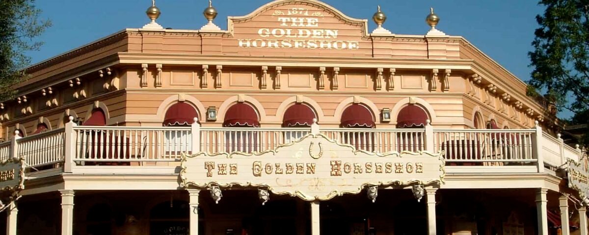 Vue frontale du bâtiment Golden Horseshoe Revue dans un parc d'attractions par une journée ensoleillée, avec une façade de deux étages de style occidental avec des gens marchant devant.