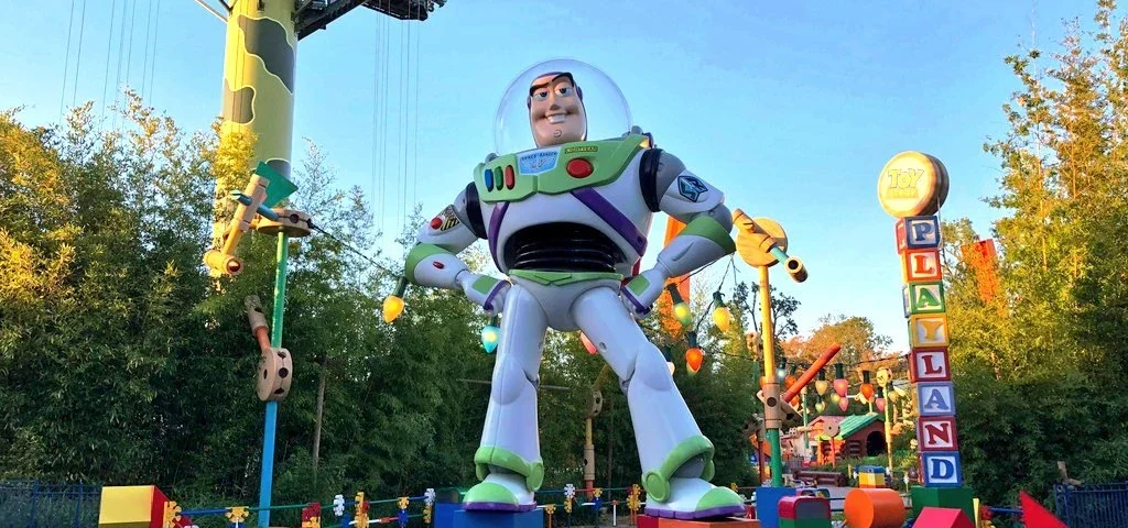Une grande statue de Buzz l'Éclair dans une pose dynamique à l'entrée de Toy Story Land au crépuscule, avec des lumières colorées et des décorations sur le thème Luxo visibles.