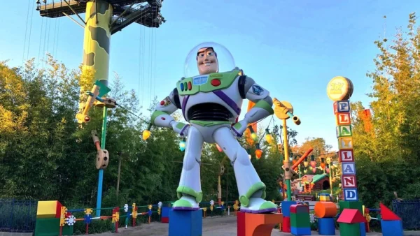 Une grande statue de Buzz l'Éclair dans une pose dynamique à l'entrée de Toy Story Land au crépuscule, avec des lumières colorées et des décorations sur le thème Luxo visibles.