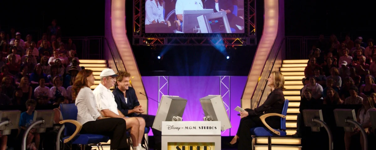 Une scène d'un jeu télévisé, "Super Soap Expedition Everest Edition", mettant en vedette deux concurrents assis en face d'un animateur à un bureau avec des moniteurs, à l'intérieur d'un studio rempli d'un public.