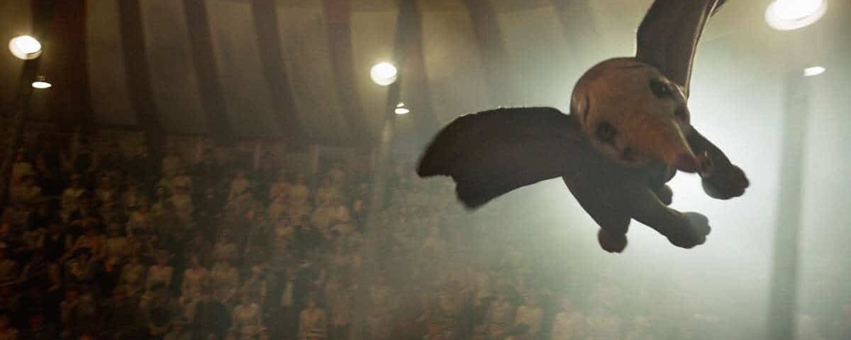 Un éléphant ressemblant à Dumbo avec de grandes ailes sombres survolant un chapiteau de cirque bondé, éclairé par des projecteurs, créant une atmosphère mystique.