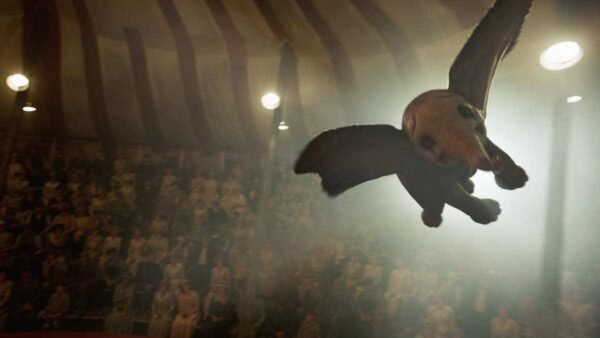 Un éléphant ressemblant à Dumbo avec de grandes ailes sombres survolant un chapiteau de cirque bondé, éclairé par des projecteurs, créant une atmosphère mystique.