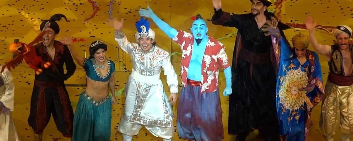 Une scène scénique vibrante mettant en vedette des acteurs habillés en personnages de « Aladdin : un spectacle musical », dont Aladdin, Jasmine et le Génie, célébrant avec un arc joyeux au milieu de confettis colorés.