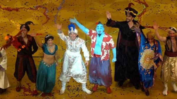 Une scène scénique vibrante mettant en vedette des acteurs habillés en personnages de « Aladdin : un spectacle musical », dont Aladdin, Jasmine et le Génie, célébrant avec un arc joyeux au milieu de confettis colorés.