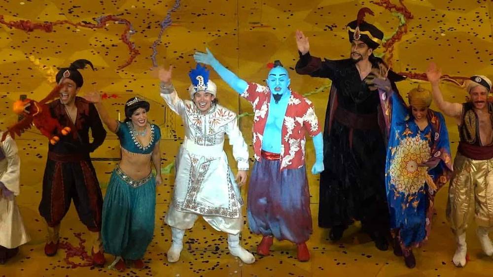 Une scène scénique vibrante mettant en vedette des acteurs habillés en personnages de « Aladdin : un spectacle musical », dont Aladdin, Jasmine et le Génie, célébrant avec un arc joyeux au milieu de confettis colorés.