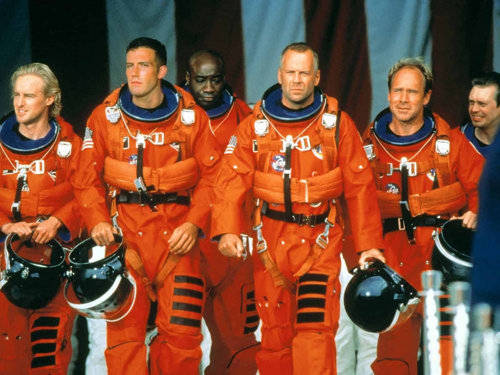 Un groupe de six astronautes vêtus de combinaisons et de casques orange vif, marchant en toute confiance avec en toile de fond un grand drapeau américain, prêts pour une mission Armageddon.