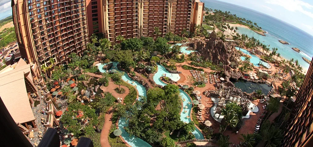 Vue aérienne du luxueux Disney Aulani Resort avec plusieurs bâtiments marron entourés d'une verdure luxuriante et de piscines sinueuses, surplombant une plage de sable fin et un océan bleu.