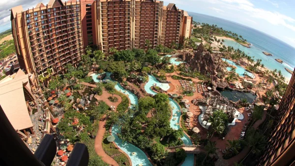 Vue aérienne du luxueux Disney Aulani Resort avec plusieurs bâtiments marron entourés d'une verdure luxuriante et de piscines sinueuses, surplombant une plage de sable fin et un océan bleu.