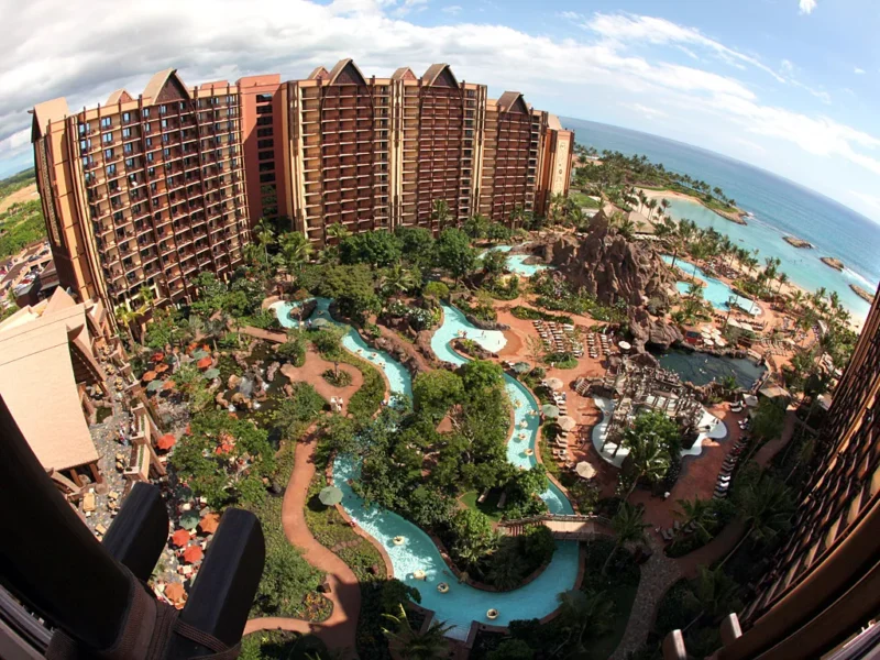 Vue aérienne du luxueux Disney Aulani Resort avec plusieurs bâtiments marron entourés d'une verdure luxuriante et de piscines sinueuses, surplombant une plage de sable fin et un océan bleu.