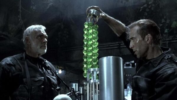 Deux hommes, dont The Rock, examinent un appareil cylindrique futuriste rempli de sphères vertes brillantes dans un cadre industriel faiblement éclairé.