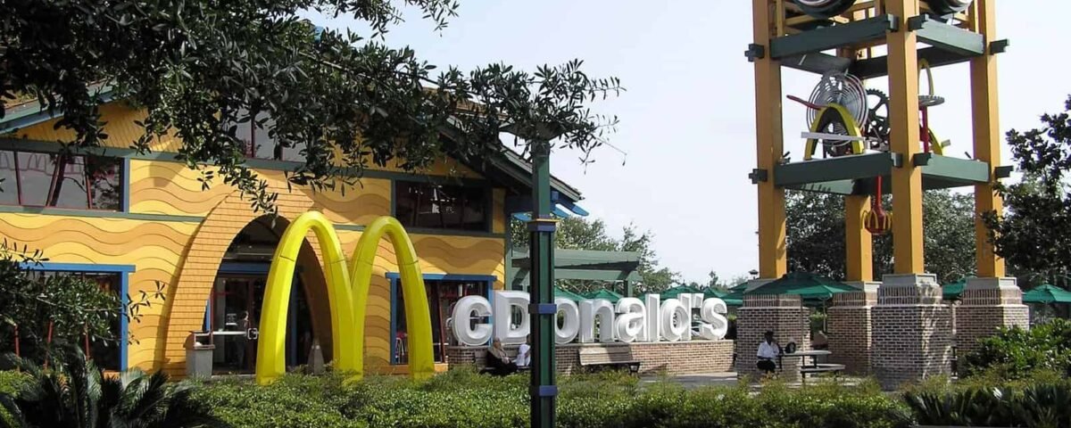 Un restaurant McDonald's au design unique avec une grande tour d'horloge à côté de ses arches dorées emblématiques, entouré d'arbres verts luxuriants et d'un ciel bleu clair.