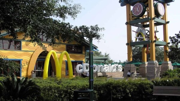 Un restaurant McDonald's au design unique avec une grande tour d'horloge à côté de ses arches dorées emblématiques, entouré d'arbres verts luxuriants et d'un ciel bleu clair.