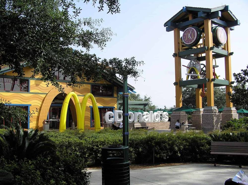 Un restaurant McDonald's au design unique avec une grande tour d'horloge à côté de ses arches dorées emblématiques, entouré d'arbres verts luxuriants et d'un ciel bleu clair.