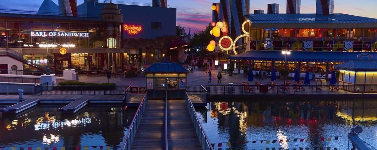 Vue en soirée d'une promenade animée en bord de mer avec des panneaux lumineux Hollywood Pictures, des restaurants et un plan d'eau calme et réfléchissant, relié par une jetée en bois sous un ciel crépusculaire.