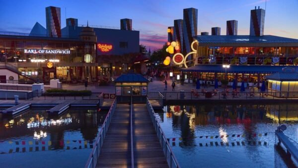 Vue en soirée d'une promenade animée en bord de mer avec des panneaux lumineux Hollywood Pictures, des restaurants et un plan d'eau calme et réfléchissant, relié par une jetée en bois sous un ciel crépusculaire.