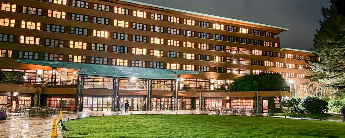 Vue nocturne d'un grand bâtiment moderne de Disneyland Paris, éclairé par un éclairage chaleureux, comprenant plusieurs étages avec de grandes fenêtres en verre et une pelouse bien entretenue au premier plan.
